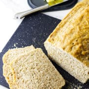A loaf of whole wheat sourdough discard sandwich bread sits on a black cutting board, with two slices cut. In the background, a plate with butter and a butter knife rests on a white surface.