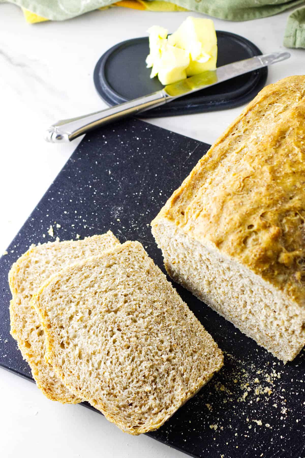A loaf of whole wheat sourdough discard sandwich bread sits on a black cutting board, with two slices cut. In the background, a plate with butter and a butter knife rests on a white surface.