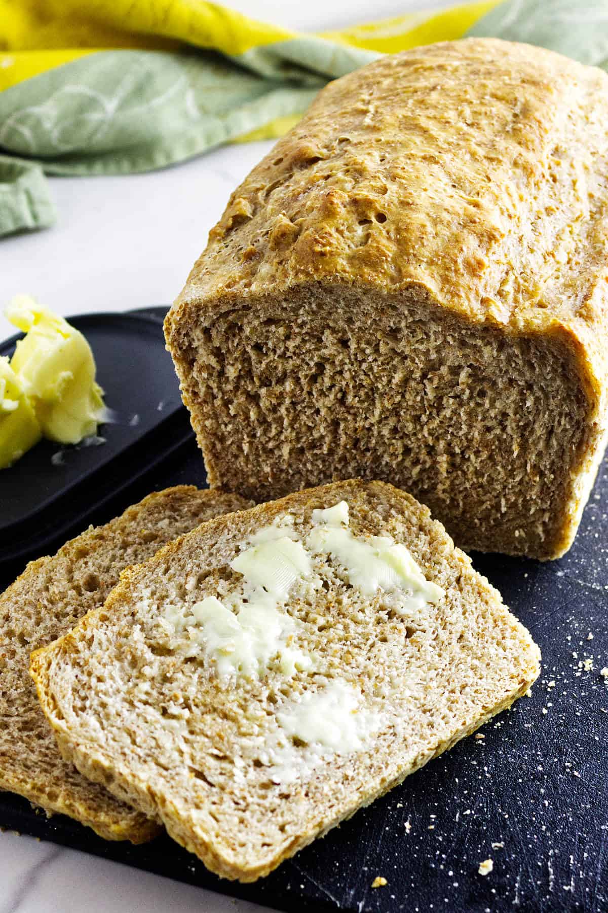 A loaf of whole wheat sourdough discard sandwich bread sits on a black cutting board, with two buttered slices in front. A dish of butter is nearby, and a yellow cloth and green towel are in the background.
