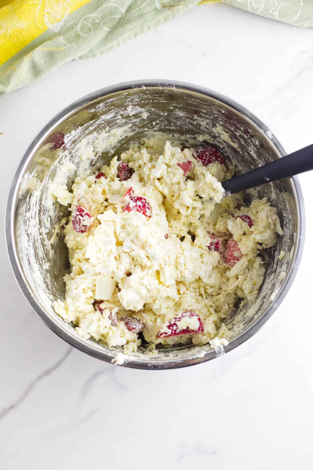 A metal mixing bowl filled with strawberry and cream cheese scone dough dotted with strawberries and white chocolate, a black spatula resting inside, all set on a white marble surface.