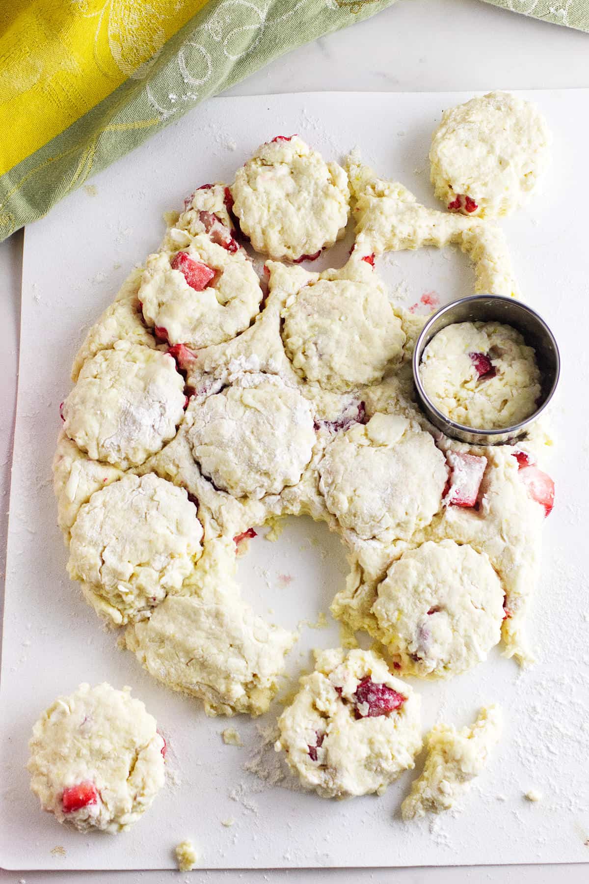 A floured surface holds biscuit dough mixed with strawberries, resembling the beginnings of strawberry and cream cheese scones. Some biscuits are cut out with a round cookie cutter, while a metal cutter rests on the uncut dough.