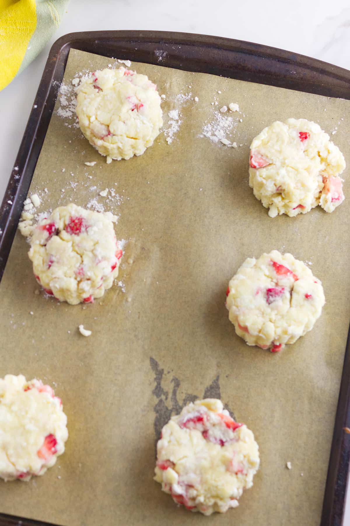 Six unbaked strawberry and cream cheese scones with visible pieces of strawberry sit on a parchment-lined baking sheet, ready to be baked. The surface around the scones is dusted with flour.