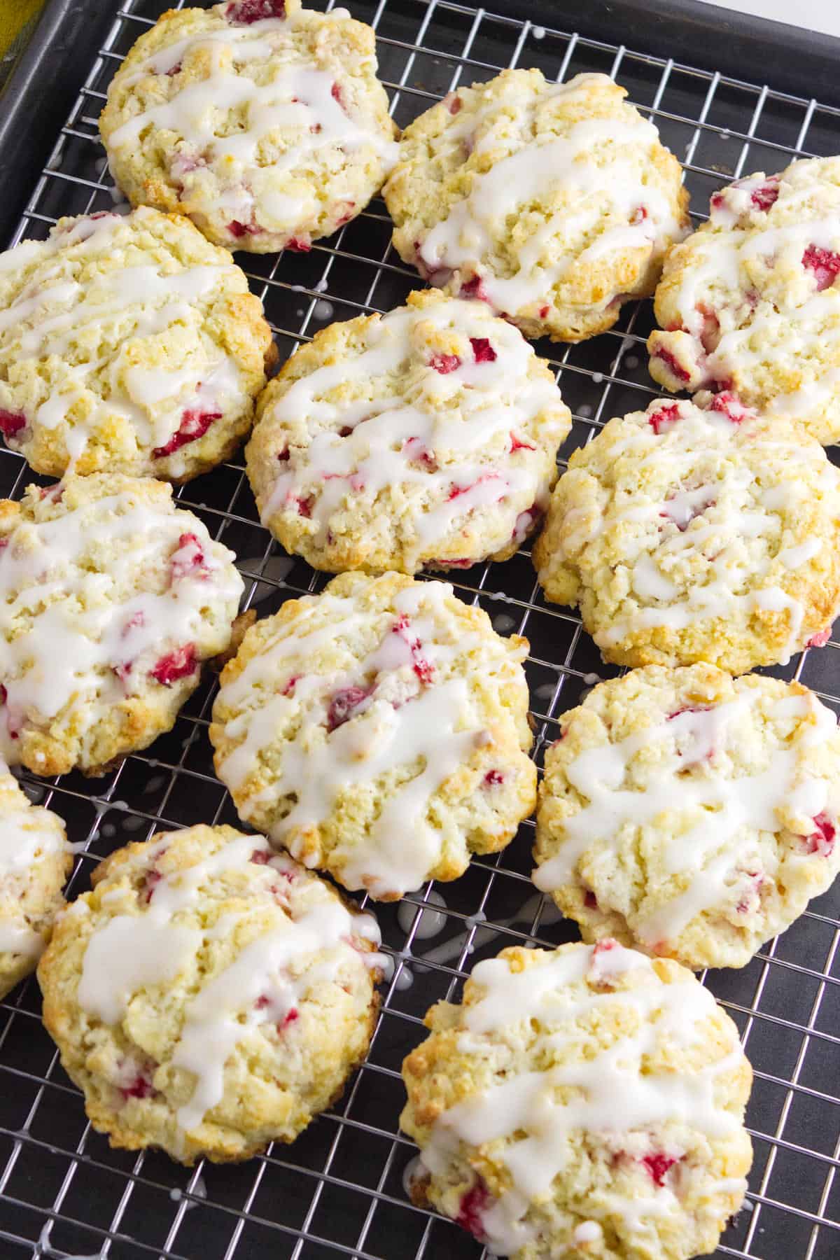 Strawberry and cream cheese scones with bits of red fruit, topped with white icing, are cooling on a wire rack placed over a baking tray.
