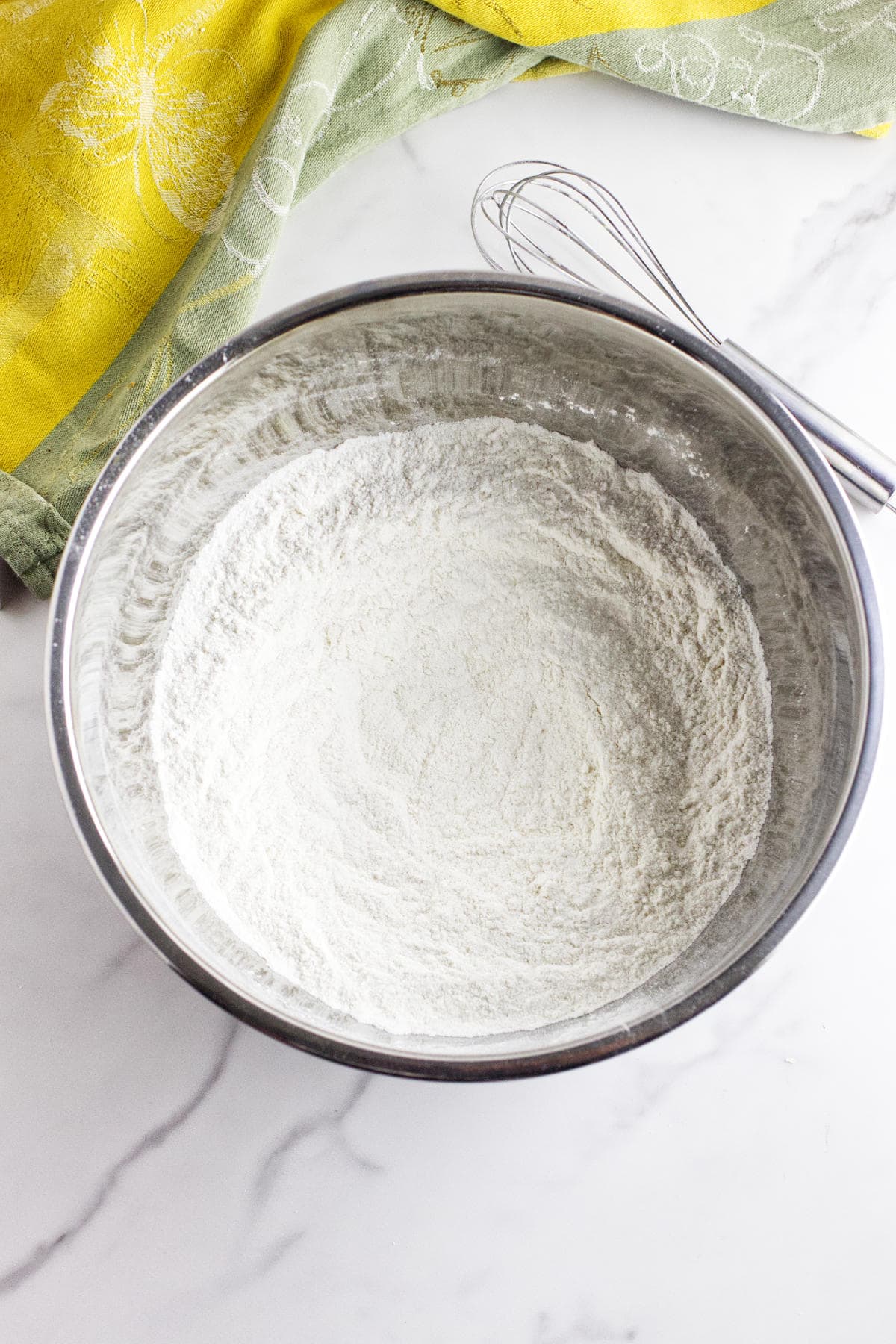 A metal mixing bowl filled with white flour sits on a marble countertop, ready for making strawberry and cream cheese scones, with a metal whisk resting on the bowl's edge. Yellow and green kitchen towels are partially visible in the background.