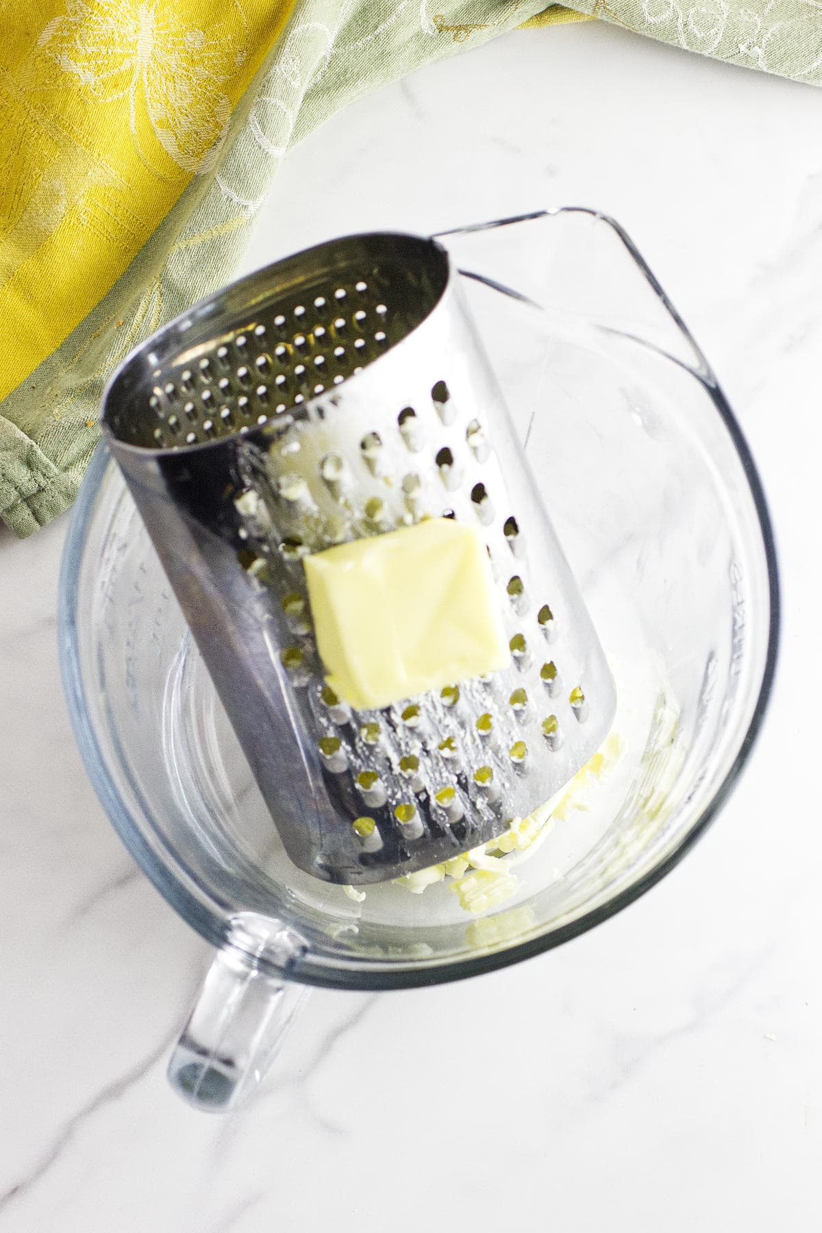 A metal box grater with a chunk of butter sits in a clear glass mixing bowl on a white countertop, ready for making strawberry and cream cheese scones, with green and yellow cloths nearby.