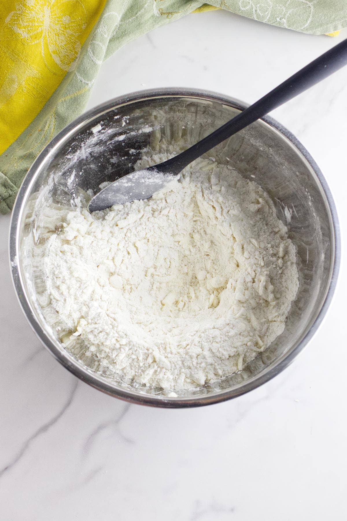 A metal mixing bowl filled with partially mixed flour and dough for strawberry and cream cheese scones sits on a white marble countertop, with a black spatula resting inside. A green and yellow cloth is in the background.