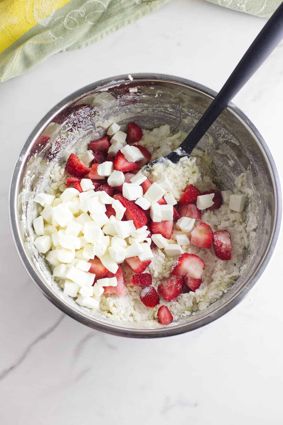 A metal mixing bowl on a white countertop holds diced strawberries and cream cheese cubes in thick batter for strawberry and cream cheese scones, with a black spoon inside. A green cloth is partially visible in the background.