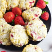 A black plate filled with freshly baked strawberry and cream cheese scones topped with white glaze, surrounded by whole strawberries. The scones have visible strawberry pieces and a crumbly texture.