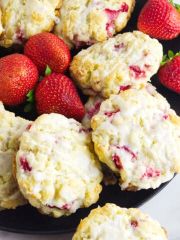 A black plate filled with freshly baked strawberry and cream cheese scones topped with white glaze, surrounded by whole strawberries. The scones have visible strawberry pieces and a crumbly texture.