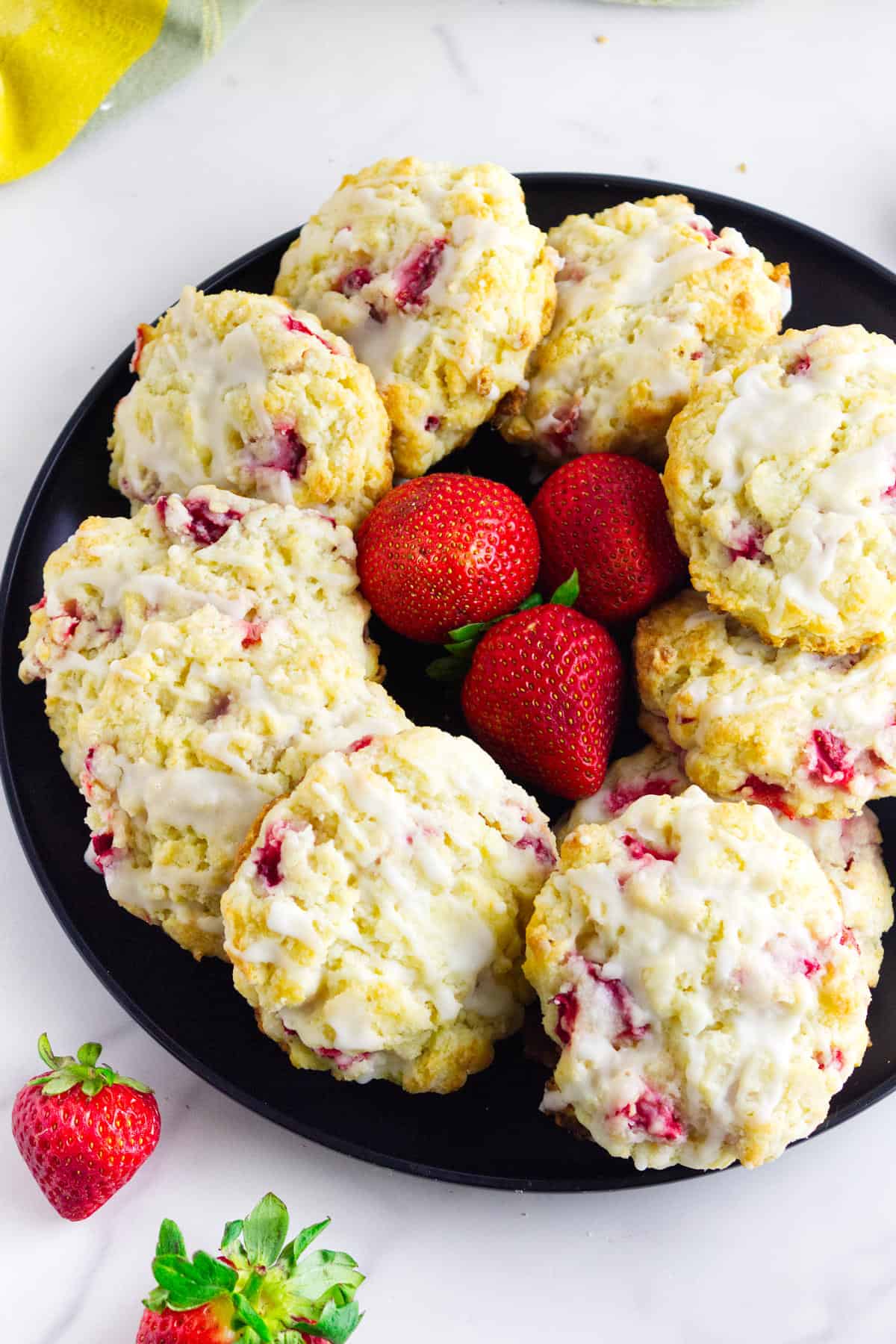 A black plate holds a circle of iced strawberry cookies with visible strawberry pieces, arranged around three fresh whole strawberries—reminiscent of strawberry and cream cheese scones. A few extra strawberries are on the white surface nearby.