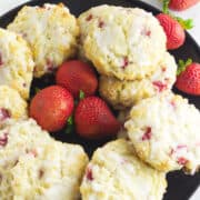 A black plate holds several glazed strawberry and cream cheese scones with visible strawberry pieces, surrounded by fresh whole strawberries on a white surface.
