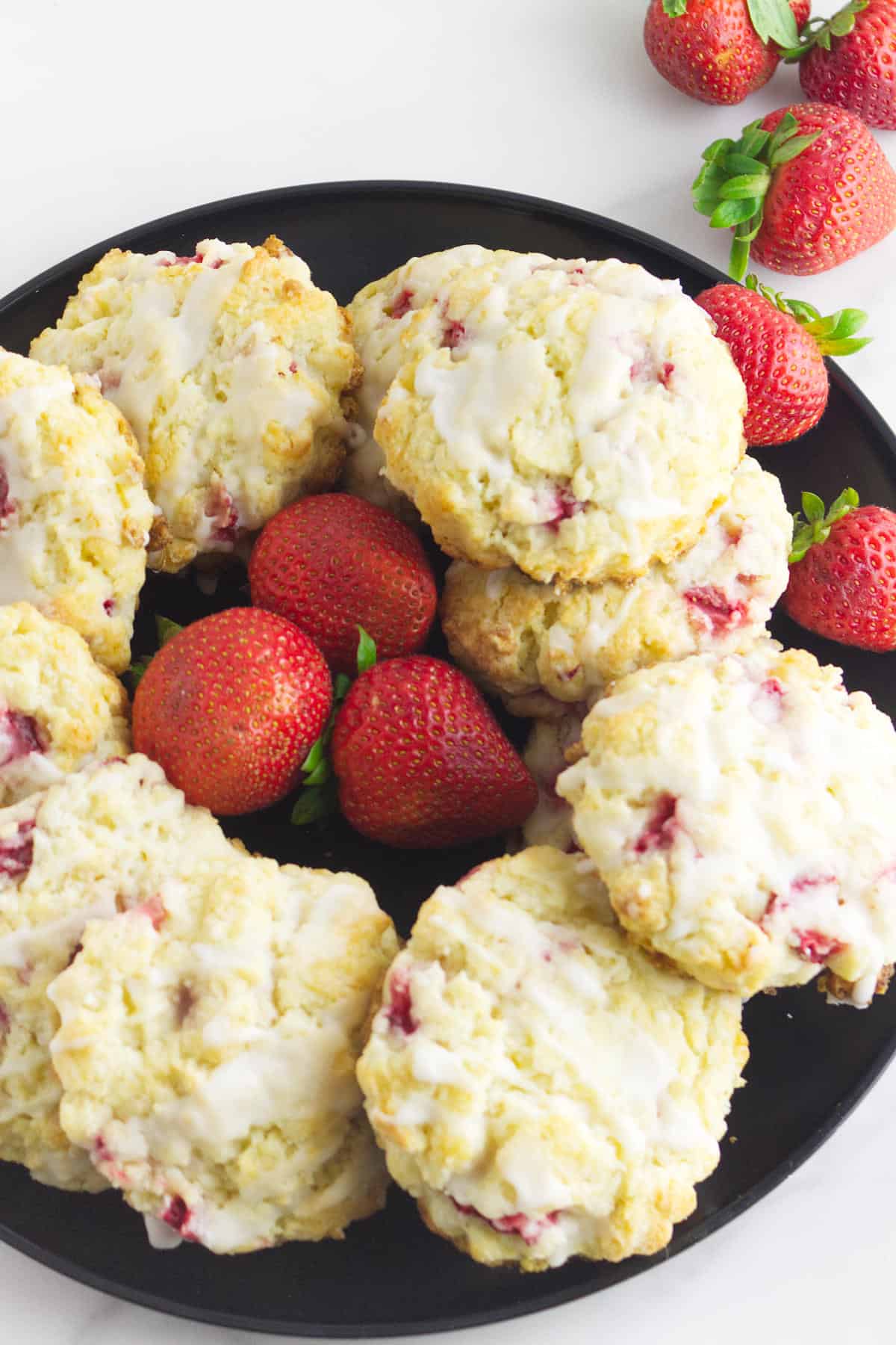 A black plate holds several glazed strawberry and cream cheese scones with visible strawberry pieces, surrounded by fresh whole strawberries on a white surface.
