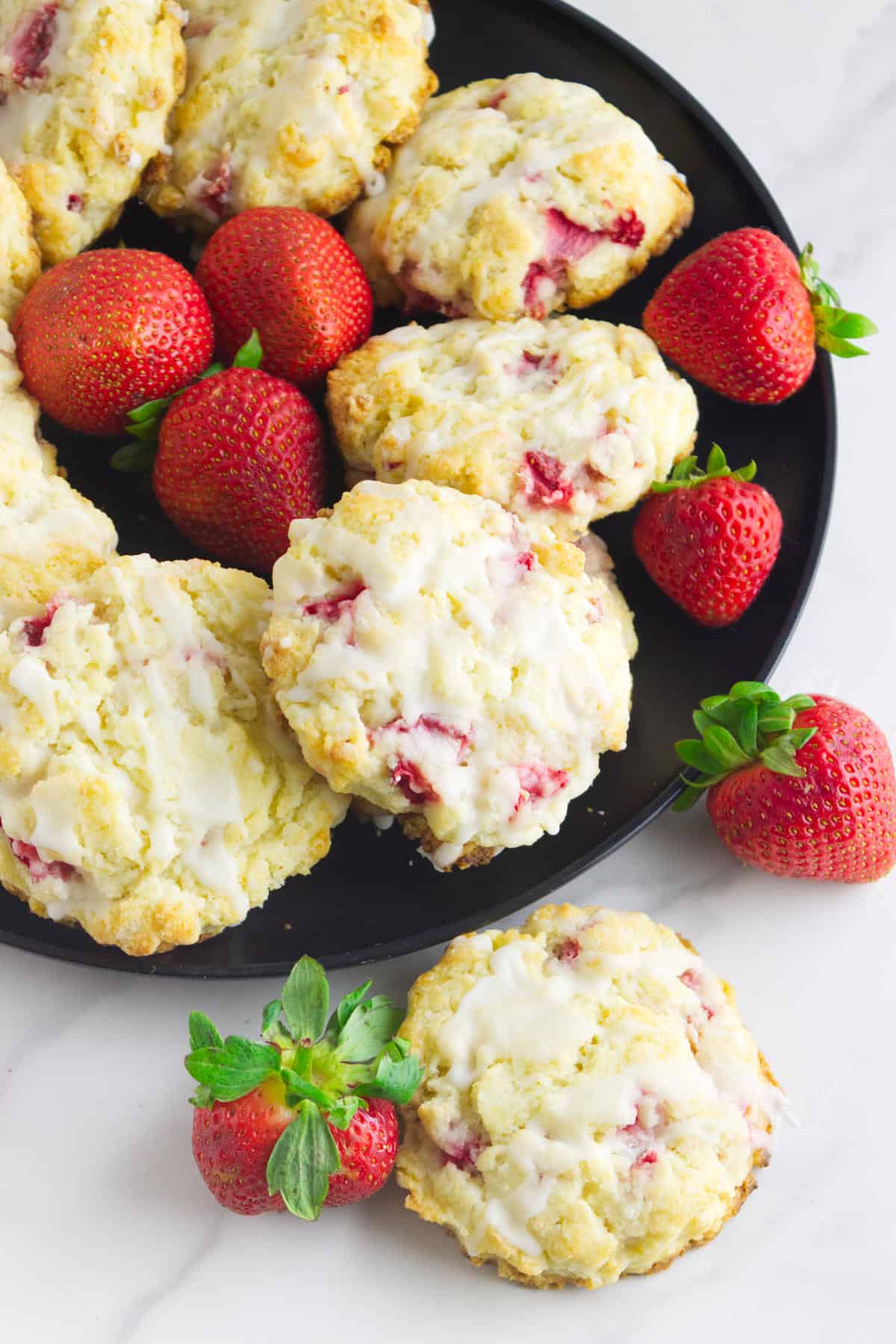 A black plate holds several glazed strawberry and cream cheese scones and fresh strawberries. Two scones and more strawberries are arranged on the white marble surface beside the plate.