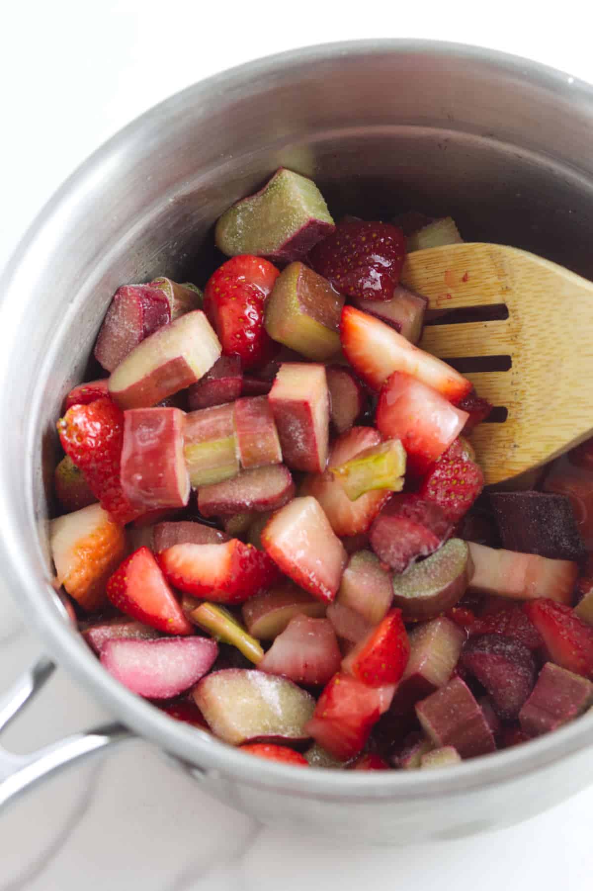 A saucepan filled with chopped strawberries and rhubarb, similar to the fruity topping for strawberry focaccia, being stirred with a wooden spatula.