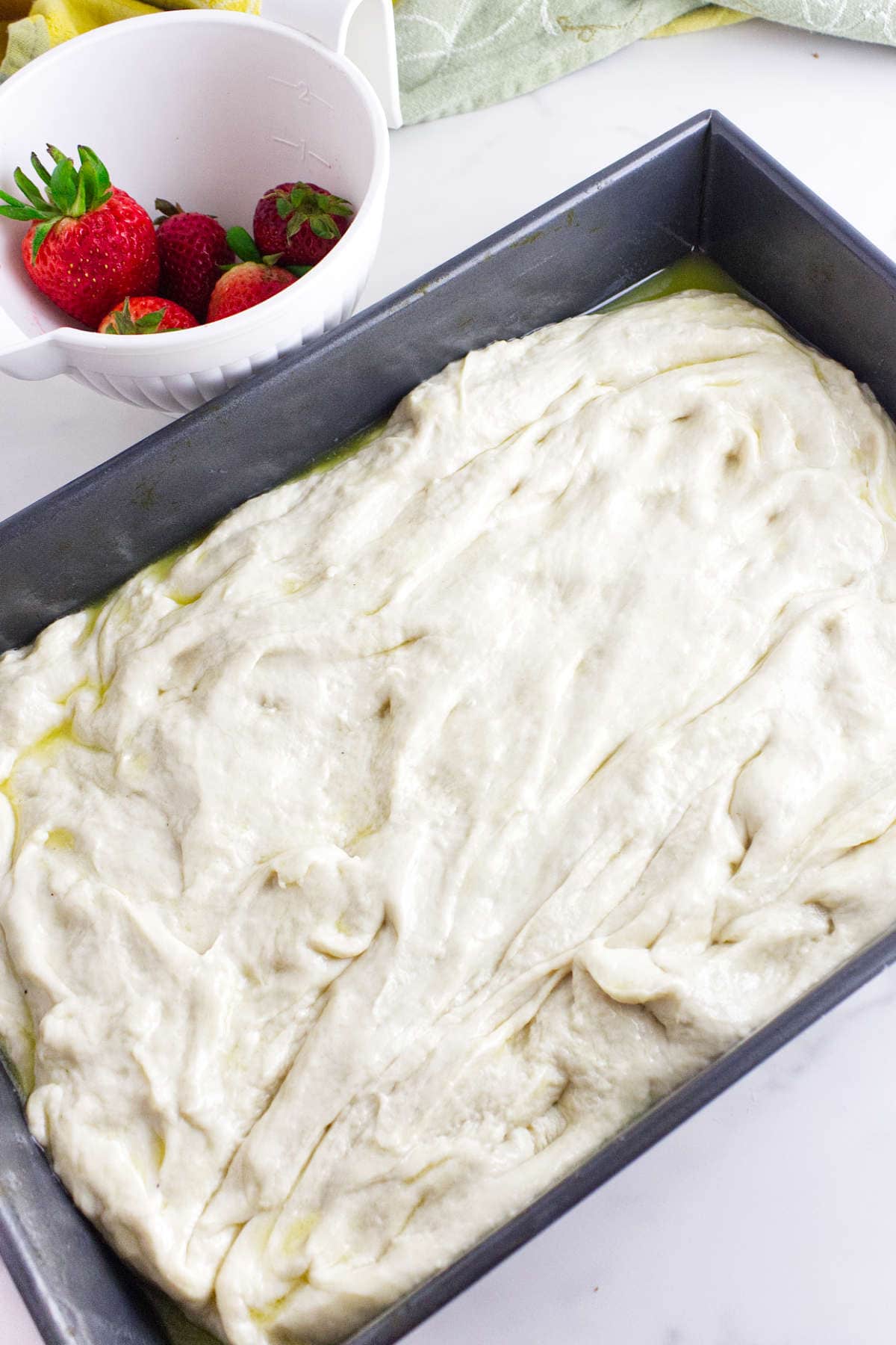A rectangular baking pan filled with raw strawberry focaccia dough sits next to a white bowl holding fresh strawberries and a couple of other berries, all on a white countertop.