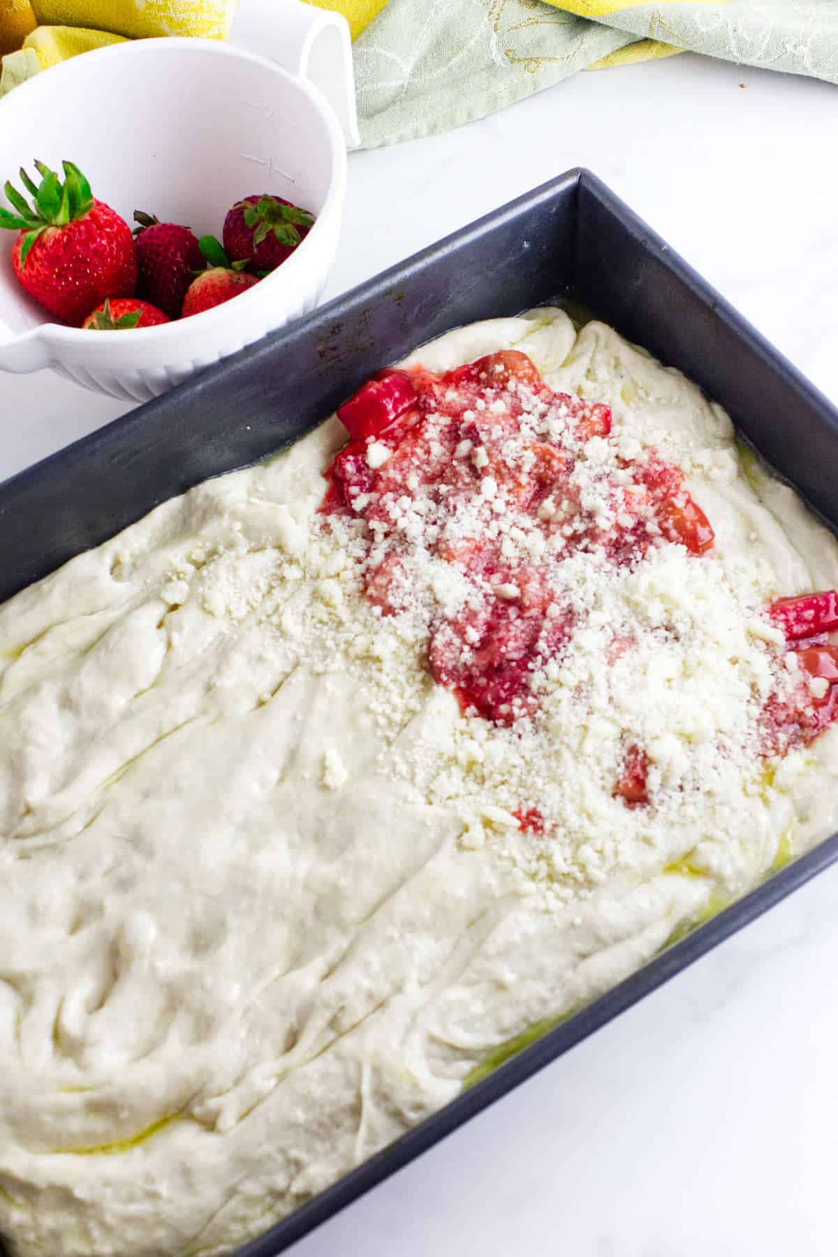 A rectangular baking pan filled with dough for strawberry focaccia, topped with chopped strawberries and sprinkled with sugar. In the background, a white bowl holds fresh strawberries.