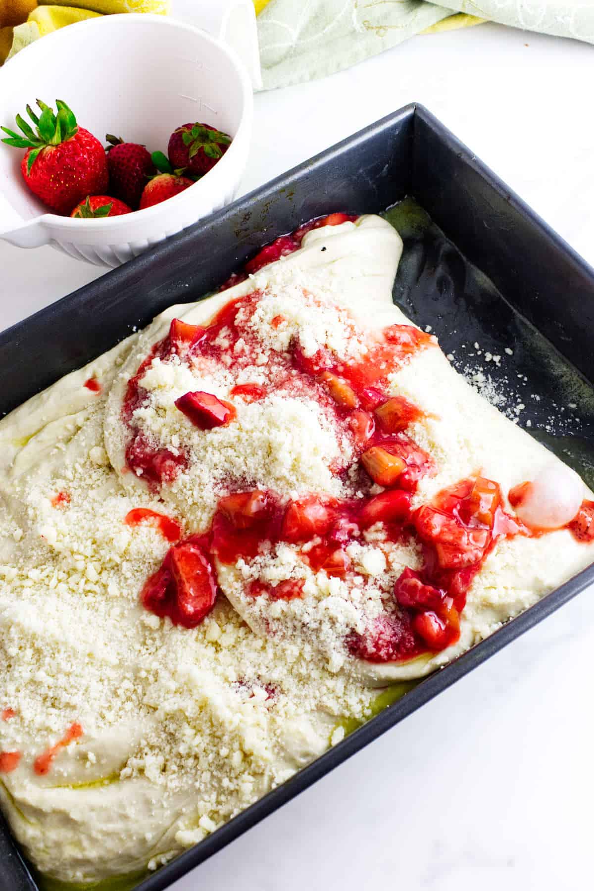 A baking pan filled with unbaked cake batter topped with strawberry compote and crumbly streusel, reminiscent of a sweet strawberry focaccia, with a bowl of fresh strawberries in the background on a white surface.