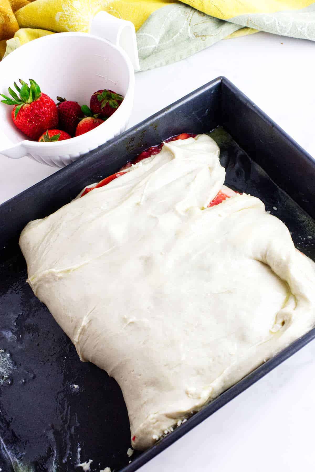 A baking pan with partially spread dough for strawberry focaccia covers sliced strawberries, next to a white bowl filled with whole strawberries on a white surface.
