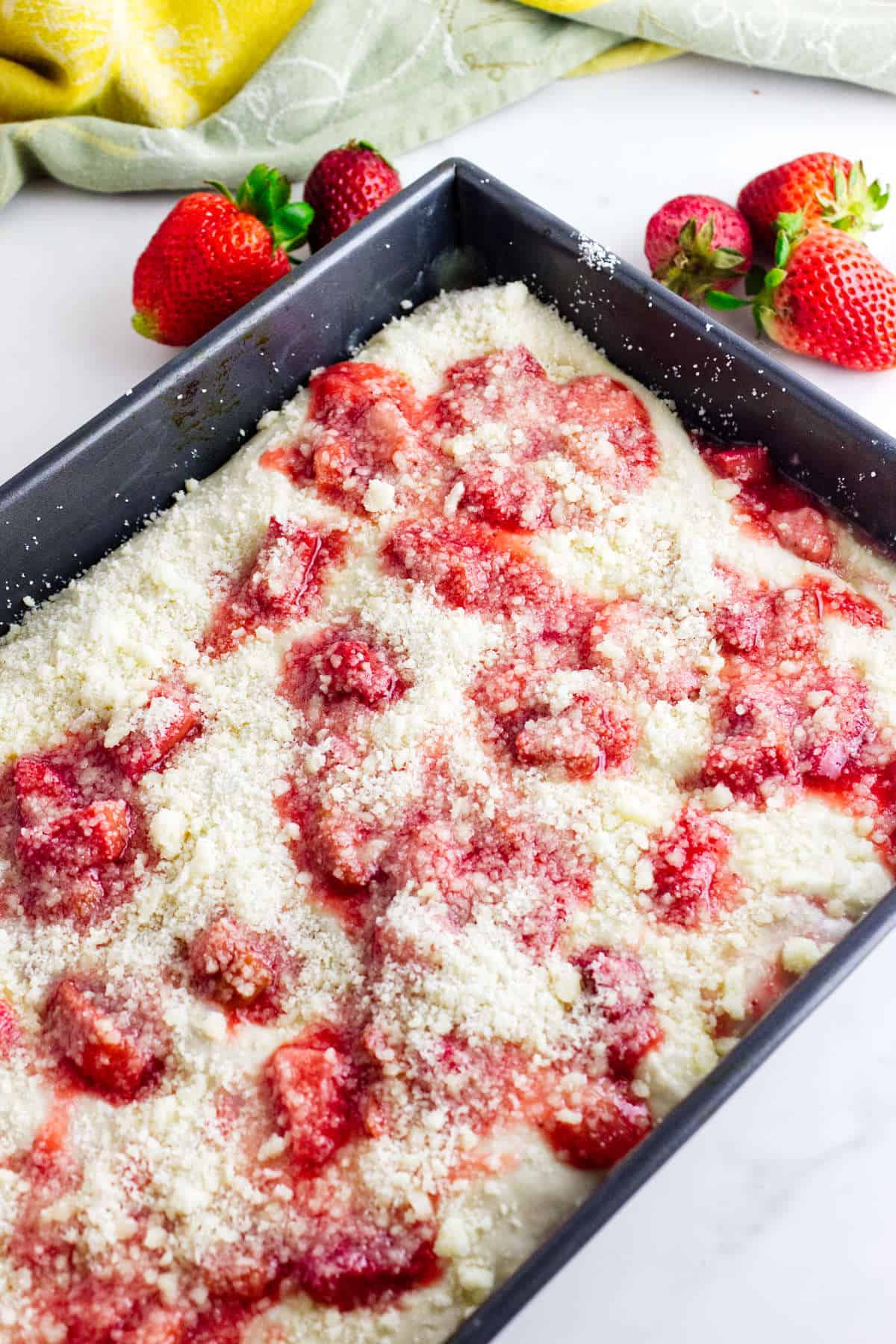 A rectangular baking pan filled with unbaked strawberry crumb cake batter, topped with strawberry pieces and crumb topping, resembles a sweet twist on strawberry focaccia. Fresh strawberries and a green cloth are in the background.