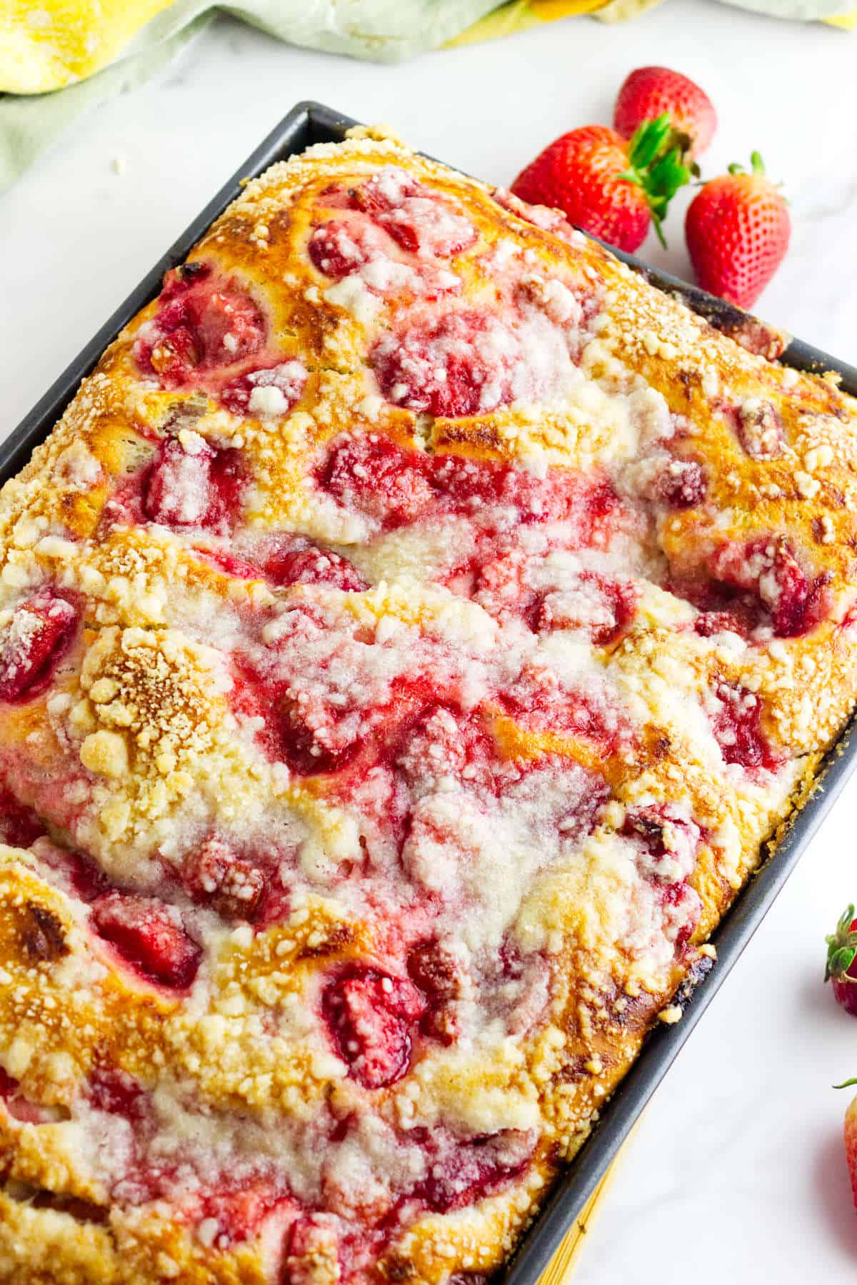 A rectangular tray of homemade strawberry coffee cake, reminiscent of strawberry focaccia, topped with sugar crumble, with fresh strawberries and a light yellow cloth in the background.
