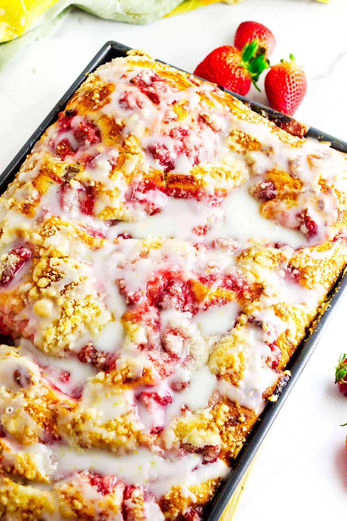 A close-up of a rectangular pan filled with glazed strawberry coffee cake. The golden cake, inspired by strawberry focaccia, is topped with pink strawberry pieces and shiny white icing. Fresh strawberries are visible in the background.