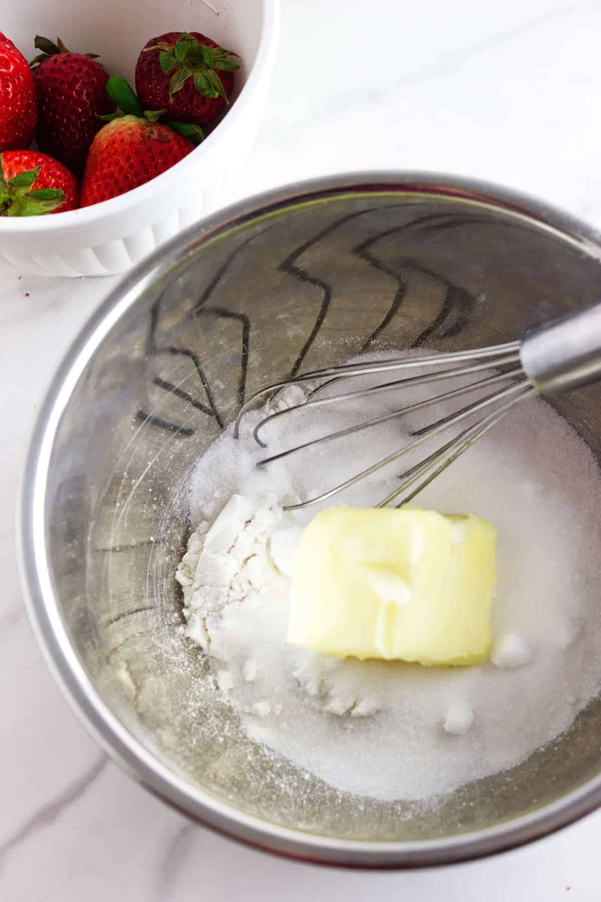 A metal mixing bowl with flour, sugar, and a stick of butter being whisked together for strawberry focaccia, next to a white bowl filled with fresh strawberries on a marble surface.