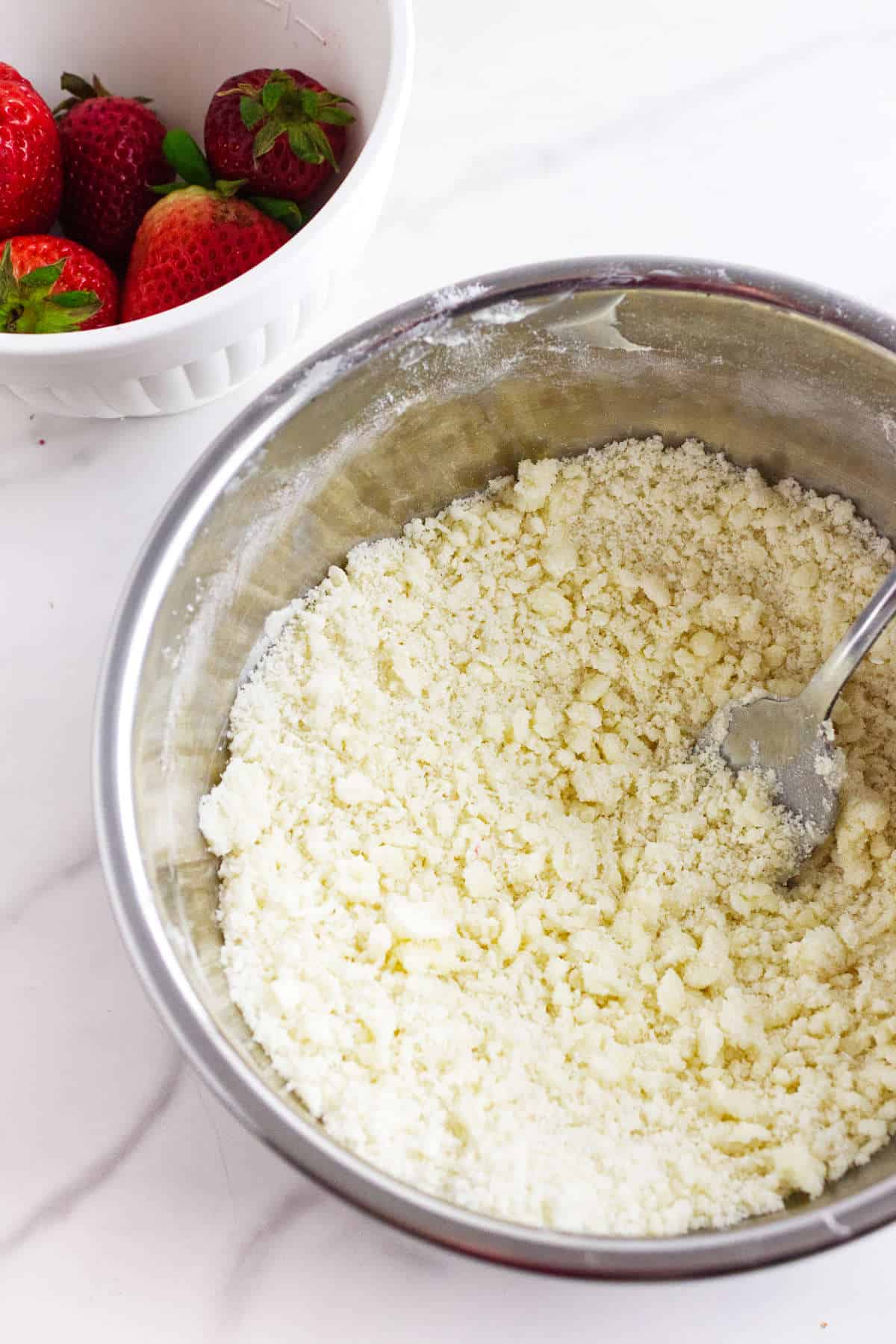 A metal bowl filled with a crumbly flour mixture and a fork sits beside a white bowl of whole strawberries on a white countertop, ready for making strawberry focaccia.