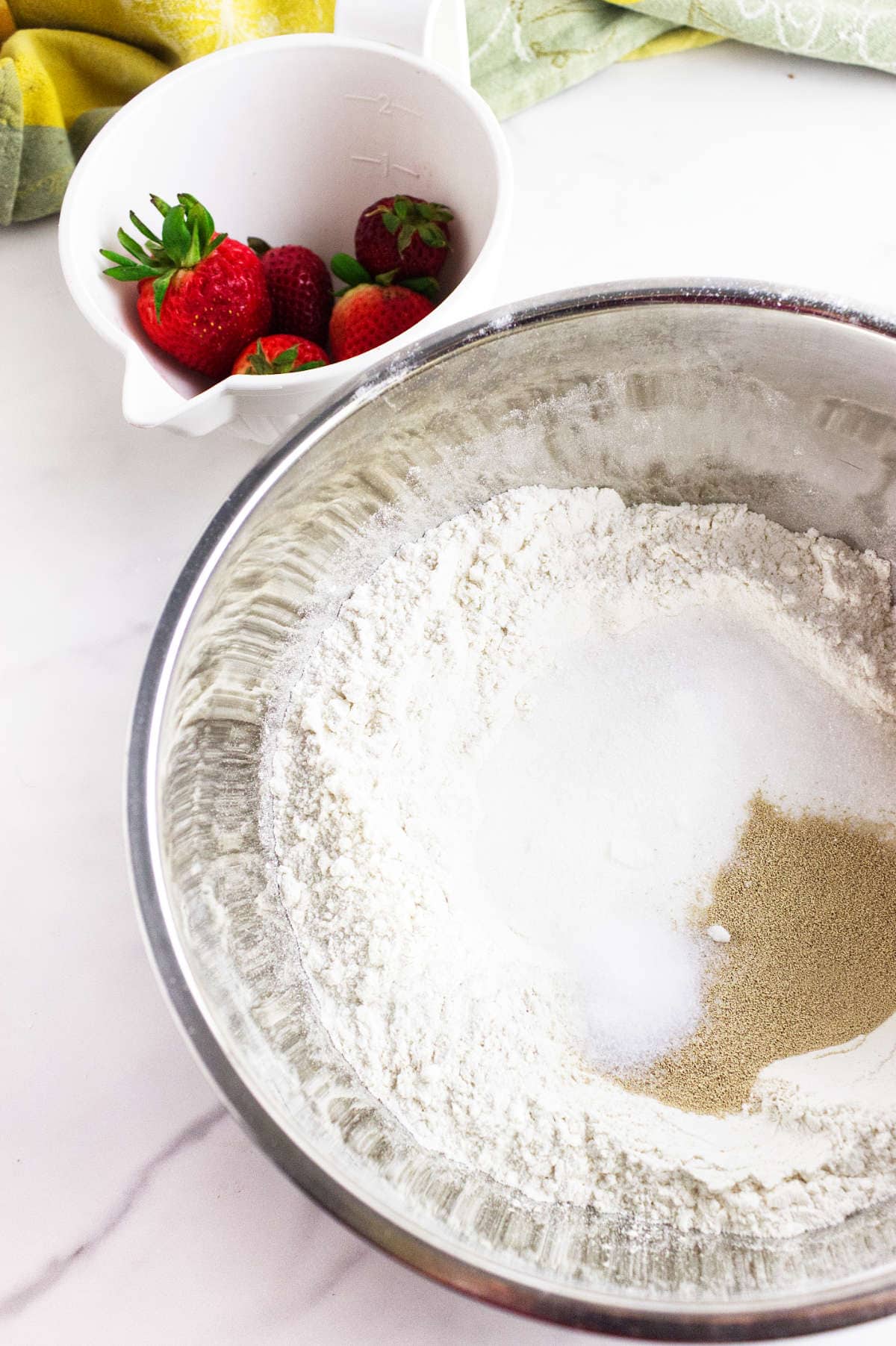 A metal mixing bowl with flour, sugar, and yeast sits on a white surface-ingredients for strawberry focaccia. Nearby is a white bowl filled with fresh strawberries, and a green and yellow cloth is partially visible in the background.
