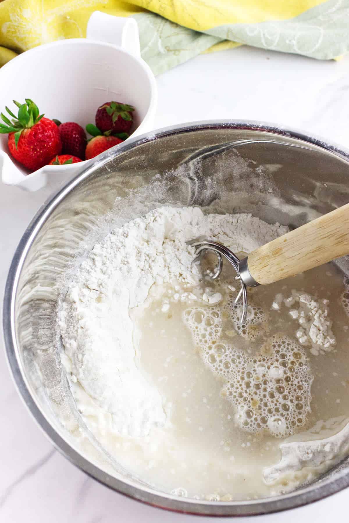 A metal mixing bowl with flour and liquid being mixed with a wooden-handled dough whisk, next to a white bowl filled with fresh strawberries-perfect for preparing homemade strawberry focaccia on a marble countertop.