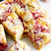 A close-up of sliced strawberry focaccia bread drizzled with white icing. The bread is golden brown with visible chunks of strawberries and a moist, crumbly texture. One slice is slightly pulled away from the loaf.