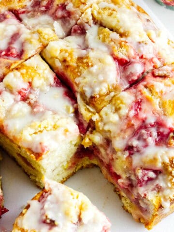 A close-up of sliced strawberry focaccia bread drizzled with white icing. The bread is golden brown with visible chunks of strawberries and a moist, crumbly texture. One slice is slightly pulled away from the loaf.