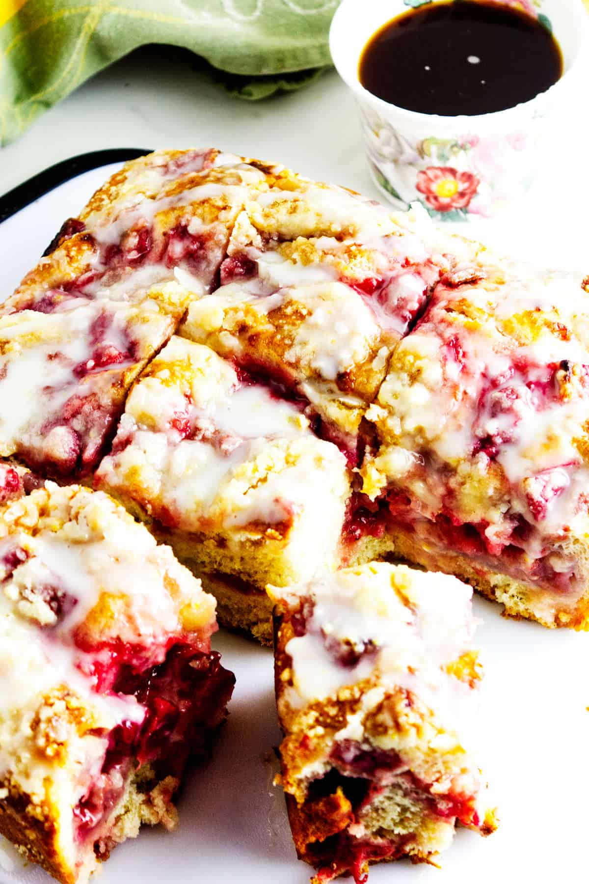 A close-up of a sliced raspberry coffee cake drizzled with white icing sits beside a cup of coffee in a floral teacup, its crumbly texture and visible raspberry filling reminiscent of a fresh strawberry focaccia.