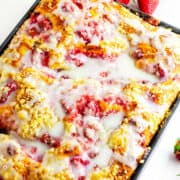 A rectangular tray of strawberry coffee cake, reminiscent of strawberry focaccia, is topped with a glossy white glaze and sits on a white surface, with whole strawberries placed nearby.