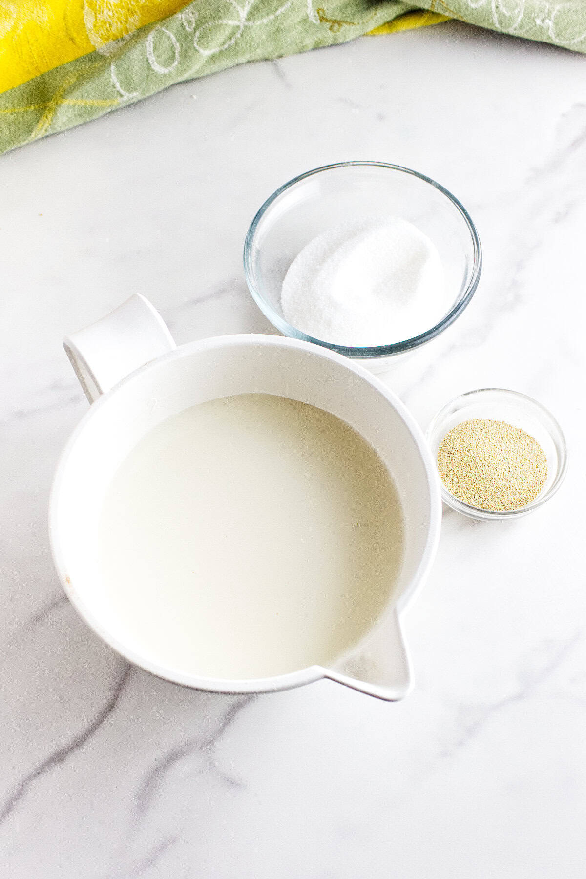 A white pitcher filled with milk sits on a marble surface next to a small bowl of sugar and a smaller bowl of dry yeast-essential ingredients for making soft Texas Roadhouse rolls. A green and yellow cloth is partially visible at the top of the image.