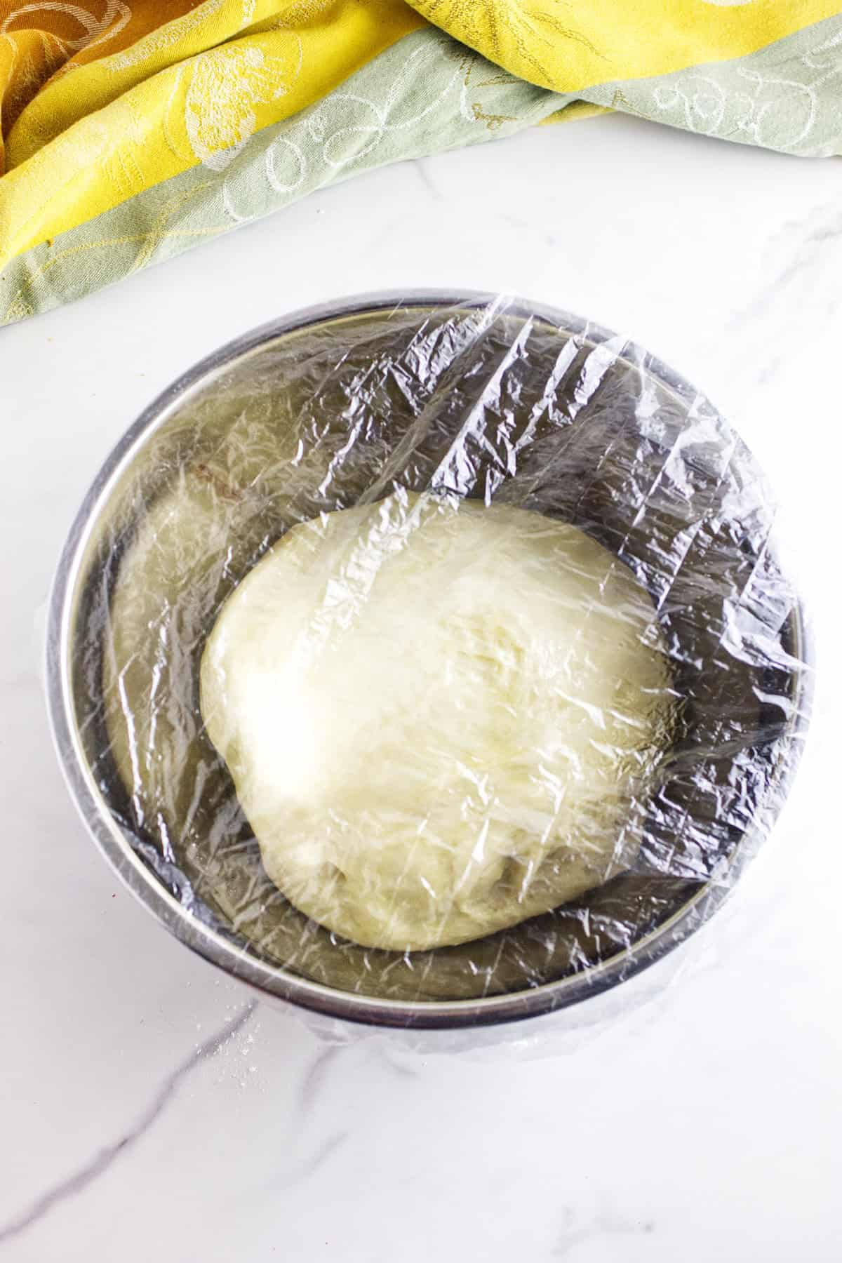 A metal bowl with dough for Texas Roadhouse rolls rising inside, covered with plastic wrap, sits on a white marble surface. A yellow and green cloth is partially visible in the upper corner.
