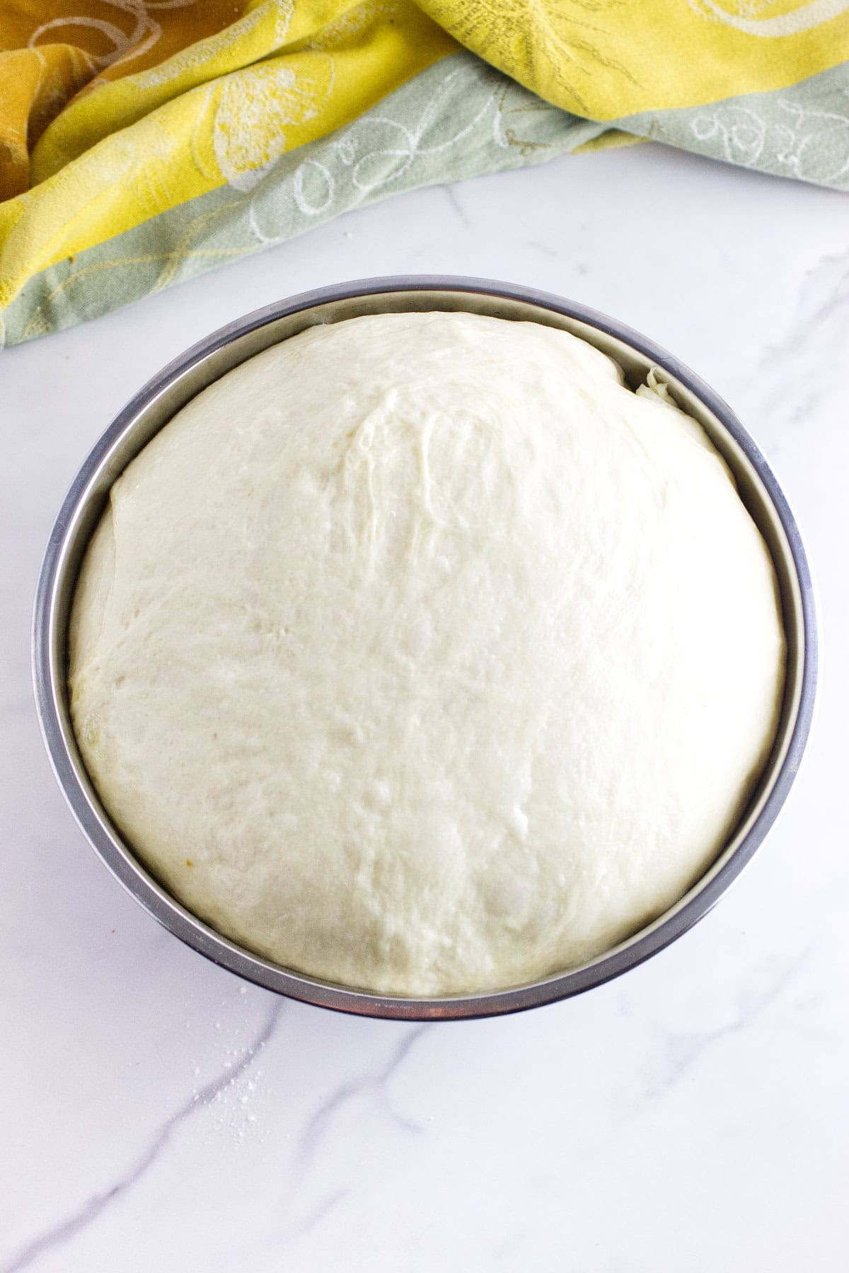A metal bowl filled with risen dough, perfect for making Texas Roadhouse rolls, sits on a white marble surface, with a yellow and green cloth partially visible in the background.