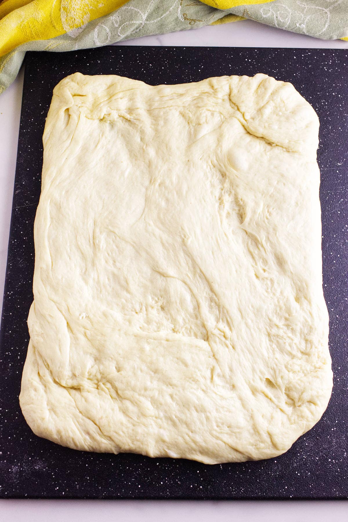 A rectangular sheet of raw, stretched dough sits on a black speckled surface, ready for baking into soft Texas Roadhouse rolls. A green and yellow cloth lies partially visible in the background.