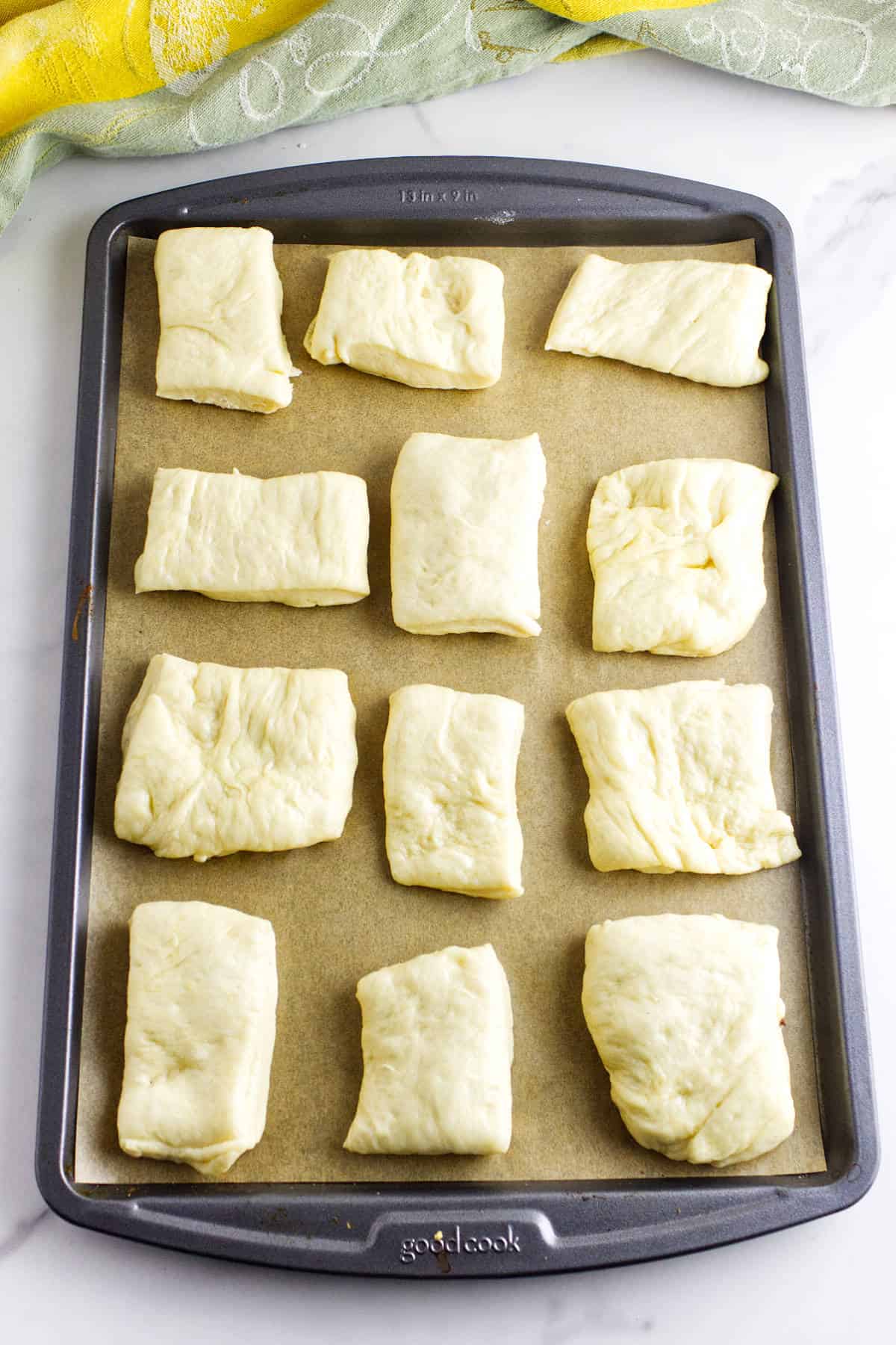 A baking sheet lined with parchment paper holds twelve unbaked, rectangular pieces of Texas Roadhouse rolls dough, spaced apart and ready for baking, on a white marble surface with a green and yellow cloth nearby.