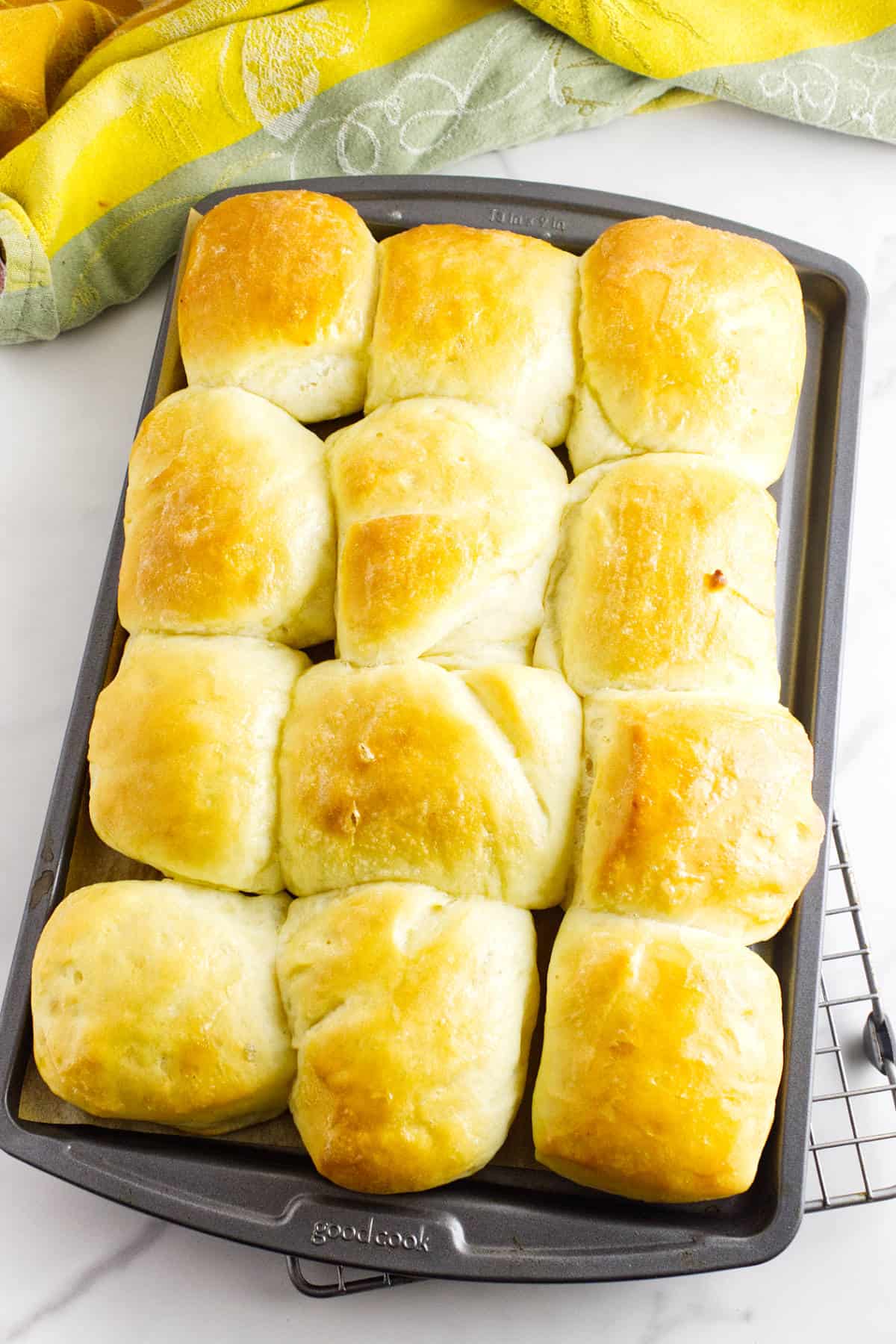 A baking tray filled with twelve golden brown Texas Roadhouse rolls, freshly baked and slightly shiny on top, rests on a cooling rack with a green and yellow cloth in the background.