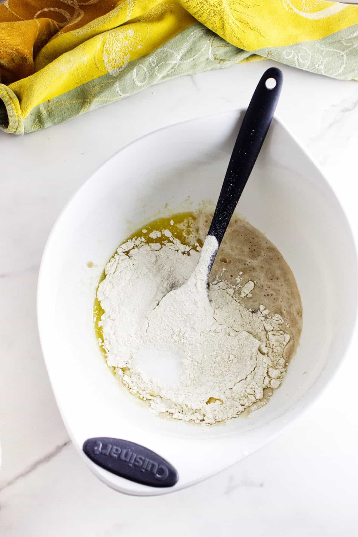 A white mixing bowl holds flour, sugar, and a wet mixture for making soft Texas Roadhouse rolls, with a black spoon resting inside. A yellow and green cloth is partially visible on a white countertop beside the bowl.