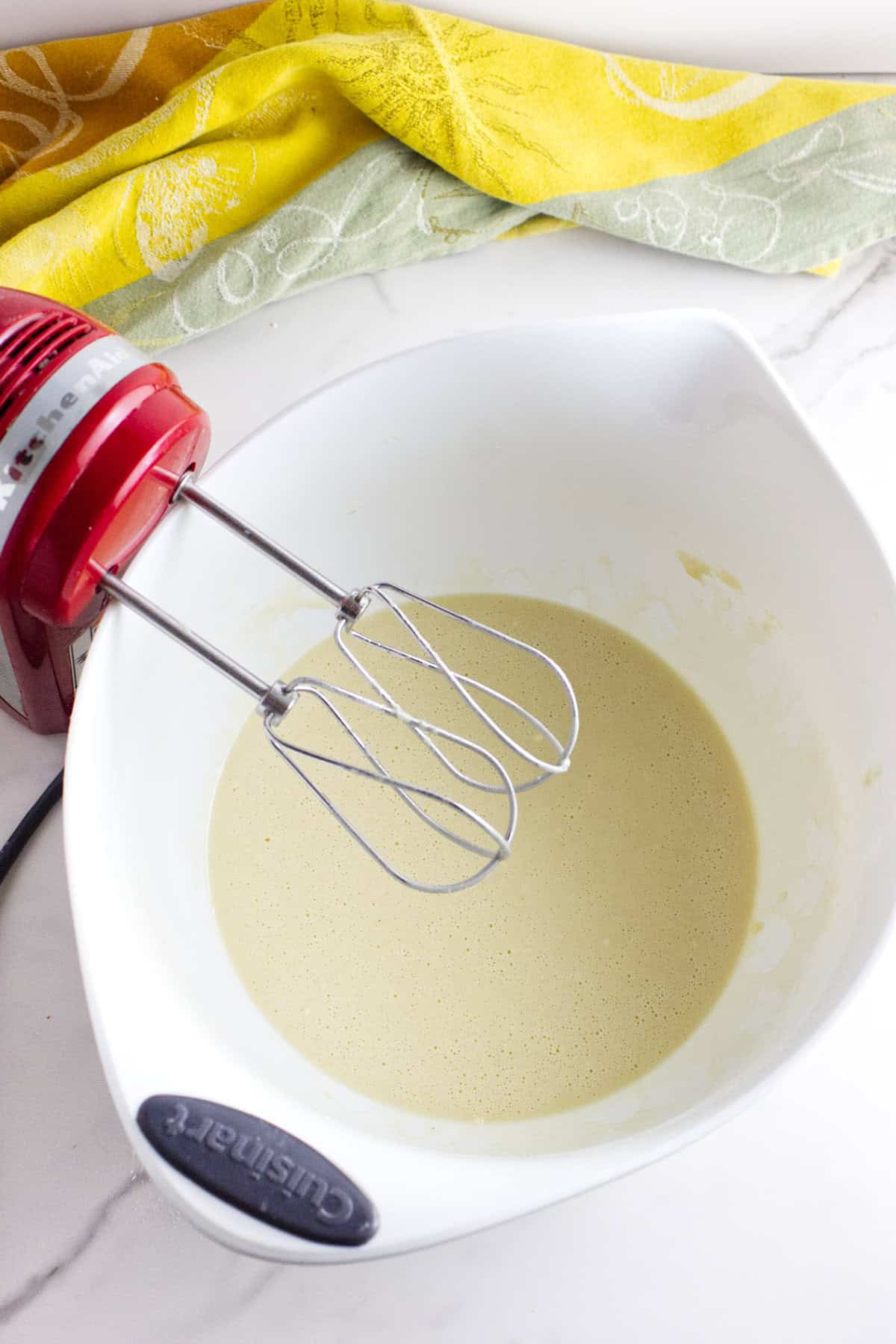 A red electric hand mixer rests in a white Cuisinart mixing bowl filled with pale, creamy batter-perfect for making Texas Roadhouse rolls. A yellow and green patterned cloth sits in the background on a marble countertop.