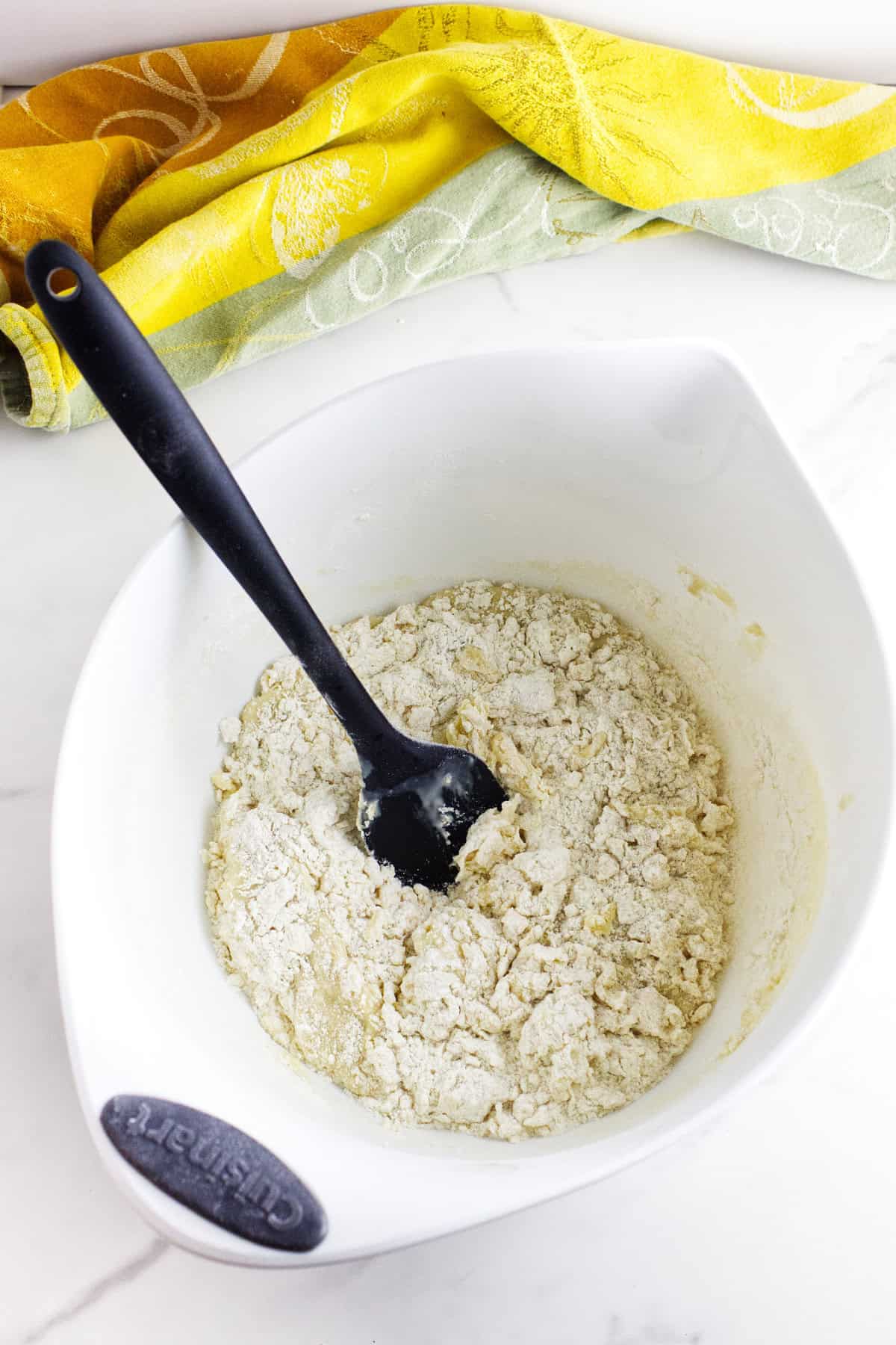 A white mixing bowl with partially mixed dough for Texas Roadhouse rolls and a black spatula inside, sits on a white surface near a yellow and green patterned cloth.