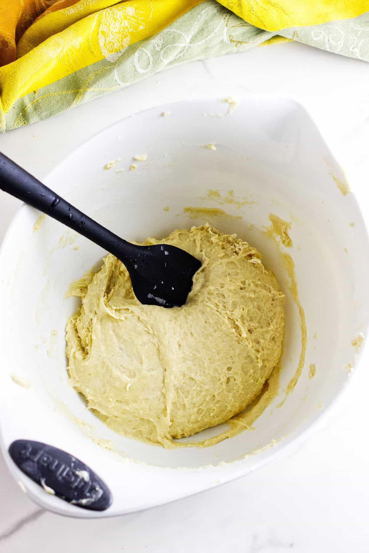 A white mixing bowl containing pale, risen dough for Texas Roadhouse rolls is being stirred with a black spatula. In the background, a green and yellow cloth is partially visible.