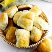 A woven basket filled with golden, fluffy Texas Roadhouse rolls sits on a white surface, with a few road house dinner rolls scattered nearby. A green and yellow cloth is partially visible in the background.