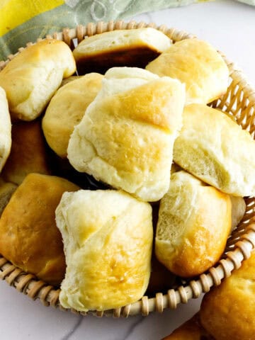 A woven basket filled with golden, fluffy Texas Roadhouse rolls sits on a white surface, with a few road house dinner rolls scattered nearby. A green and yellow cloth is partially visible in the background.