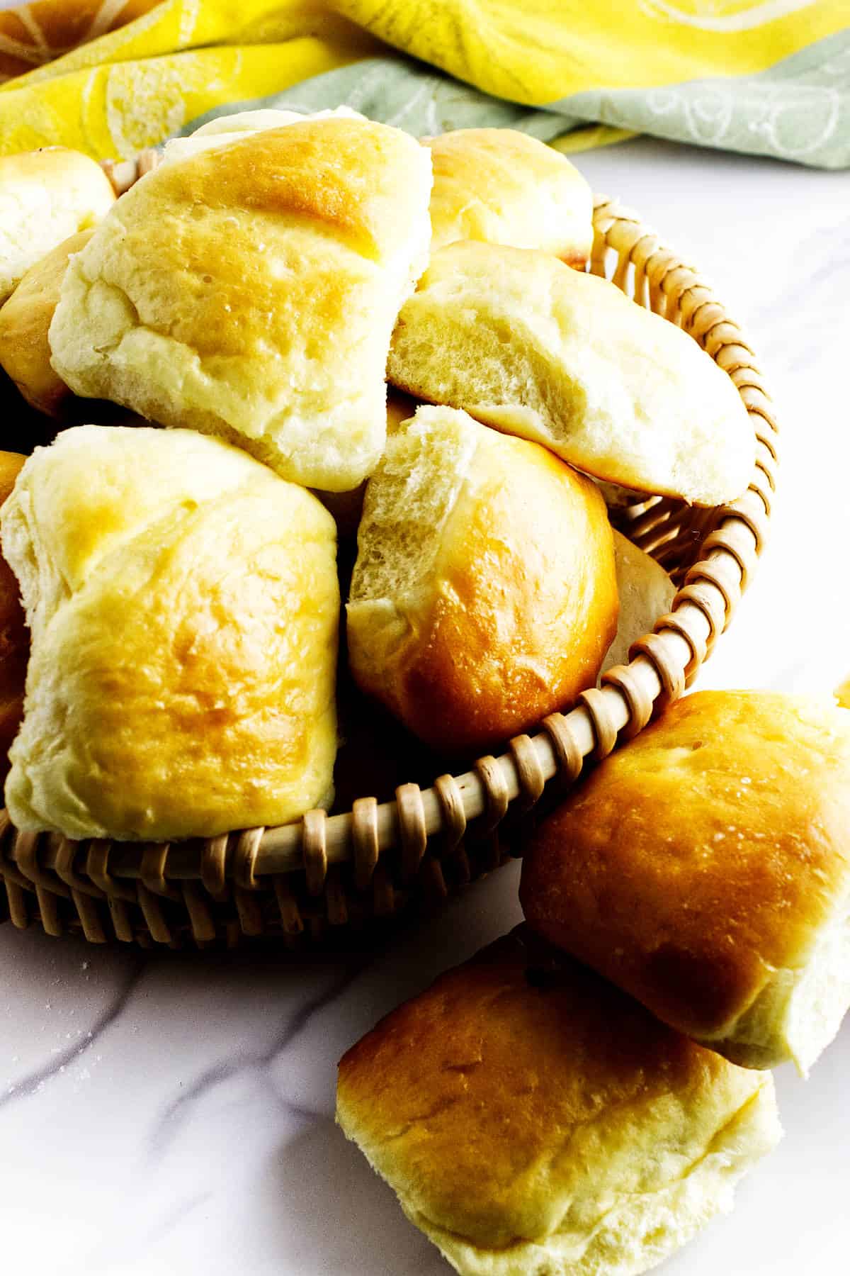 A basket filled with golden, fluffy Texas Roadhouse rolls sits on a white surface, with a few rolls placed around the basket. A yellow cloth is partially visible in the background.