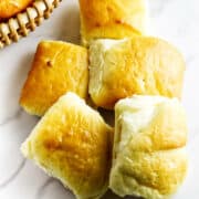 Five golden, fluffy Texas Roadhouse rolls are arranged on a white marble surface, with part of a woven basket containing more road house dinner rolls visible in the upper left corner.