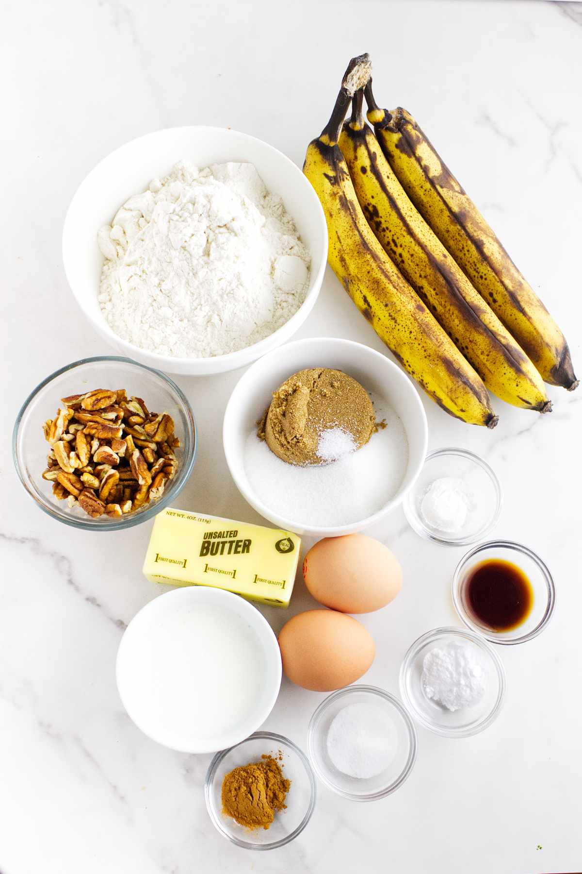 A marble countertop displays the ingredients for bread machine banana bread: ripe bananas, flour, chopped pecans, sugars, eggs, butter, milk, cinnamon, salt, baking soda and powder, and vanilla extract arranged in bowls.