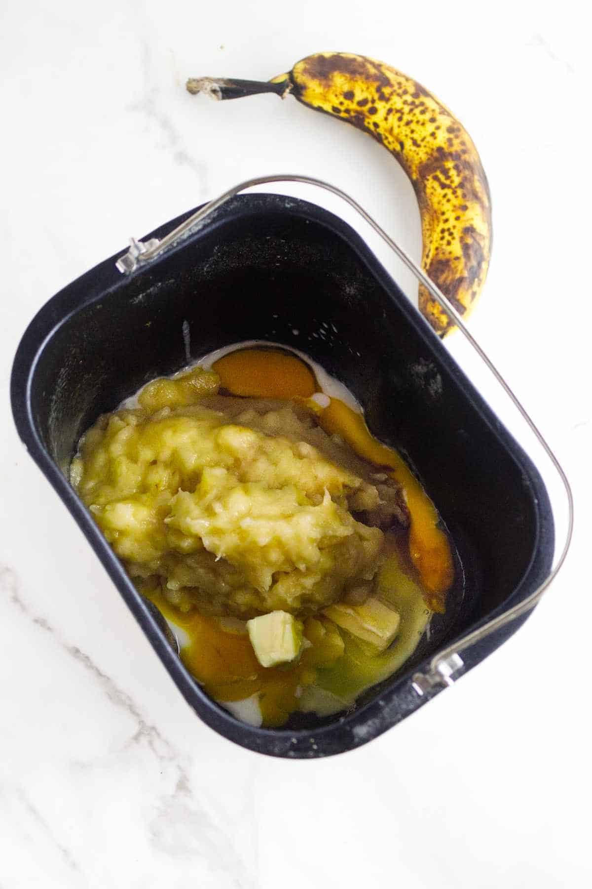 A black container holds mashed bananas, melted butter, and eggs-ingredients for delicious bread machine banana bread. A ripe, spotted banana rests on a white marble surface beside the container.