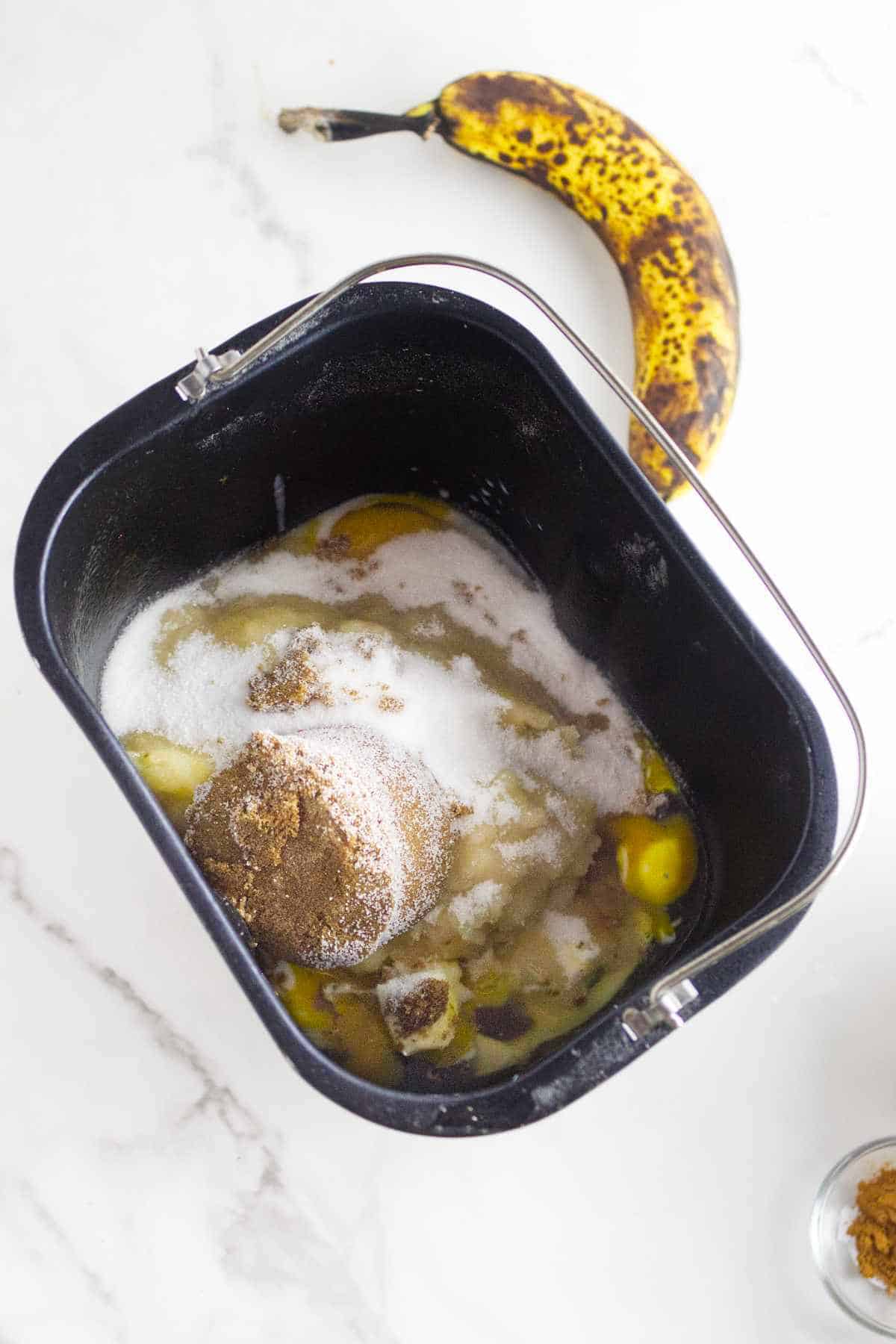 A bread machine pan filled with sugar, brown sugar, eggs, and other ingredients sits on a white surface, ready to make delicious bread machine banana bread next to an overripe banana with brown spots.
